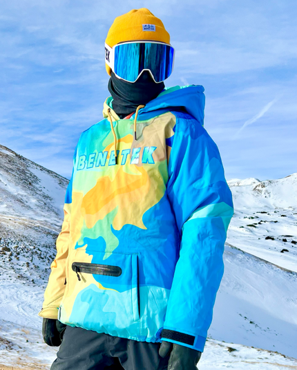 A person wearing a Benetek branded waterproof hoodie with a colorful design, including blue, yellow, and green, with a hood, sunglasses, and gloves, standing in a snowy landscape.