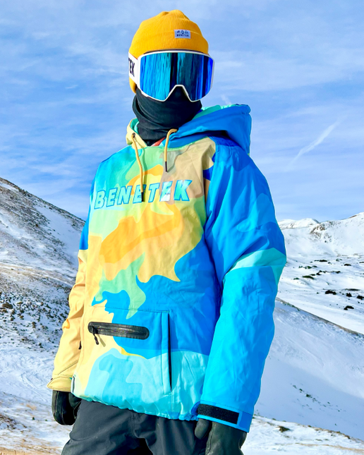 A person wearing a Benetek branded waterproof hoodie with a colorful design, including blue, yellow, and green, with a hood, sunglasses, and gloves, standing in a snowy landscape.